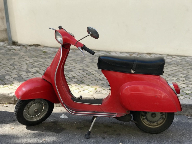 A red vintage scooter parked on a cobblestone street beside a cream wall, original photo
